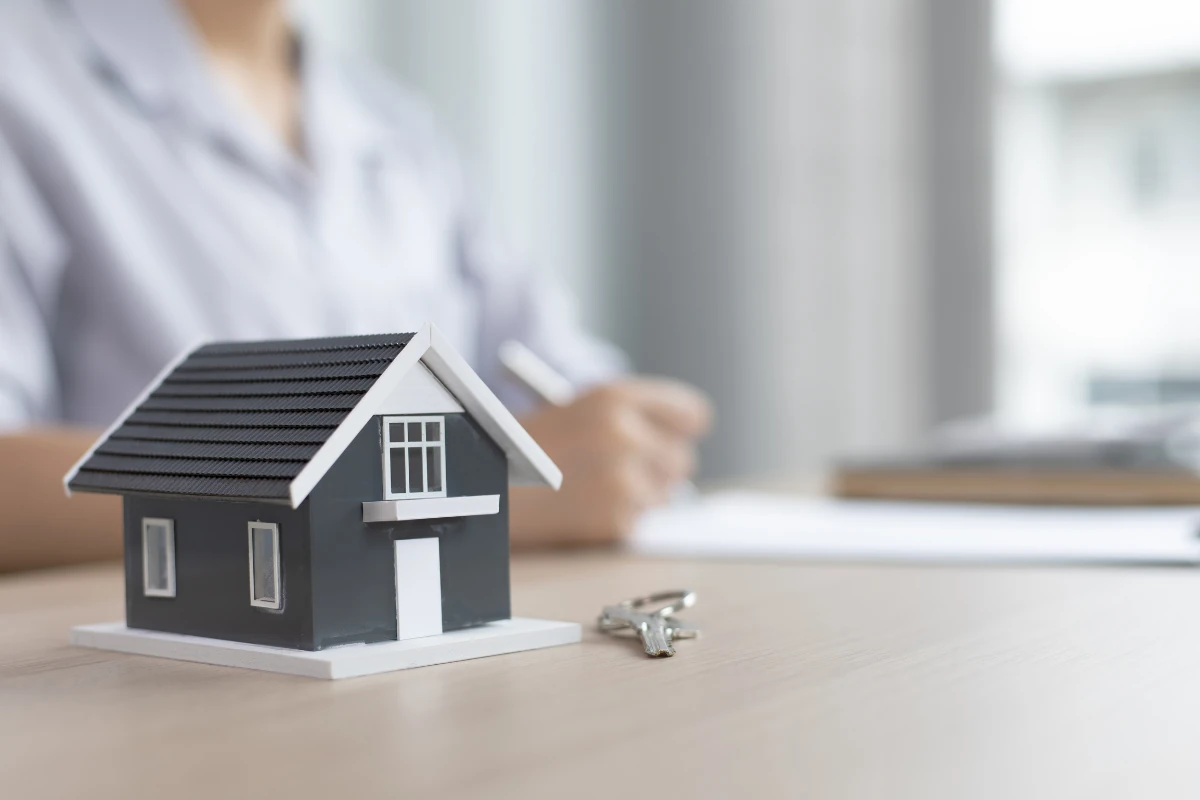Photo of a model house with realtor working in the background and a house key laying on the table